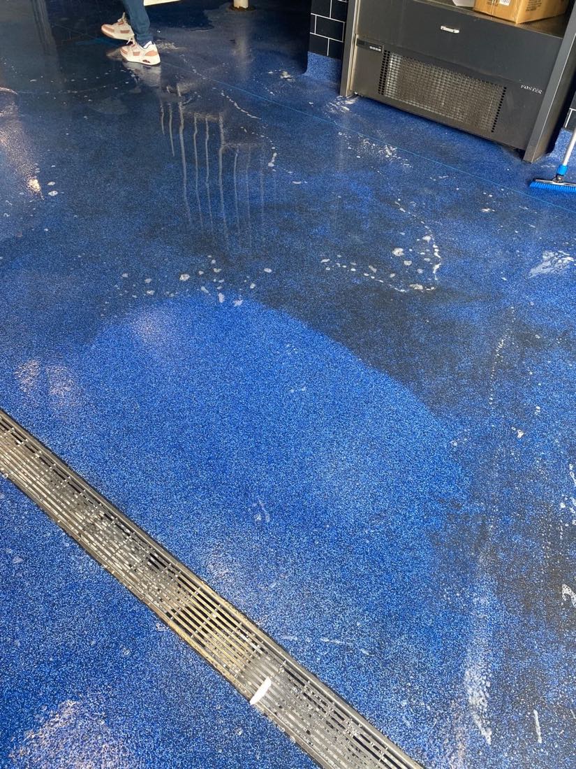 Blue safety-floor pool surround mid deep-clean — drainage channel and one section scrubbed back to a clean, even finish